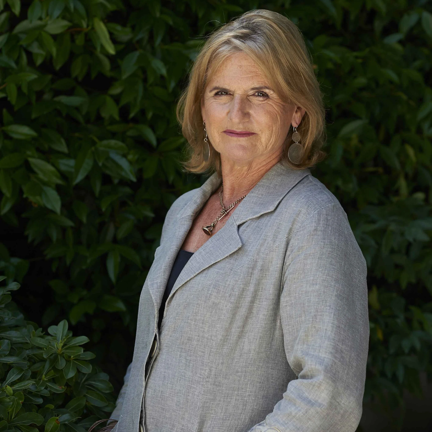 A headshot of a woman in a gray blazer with short blonde hair in a garden