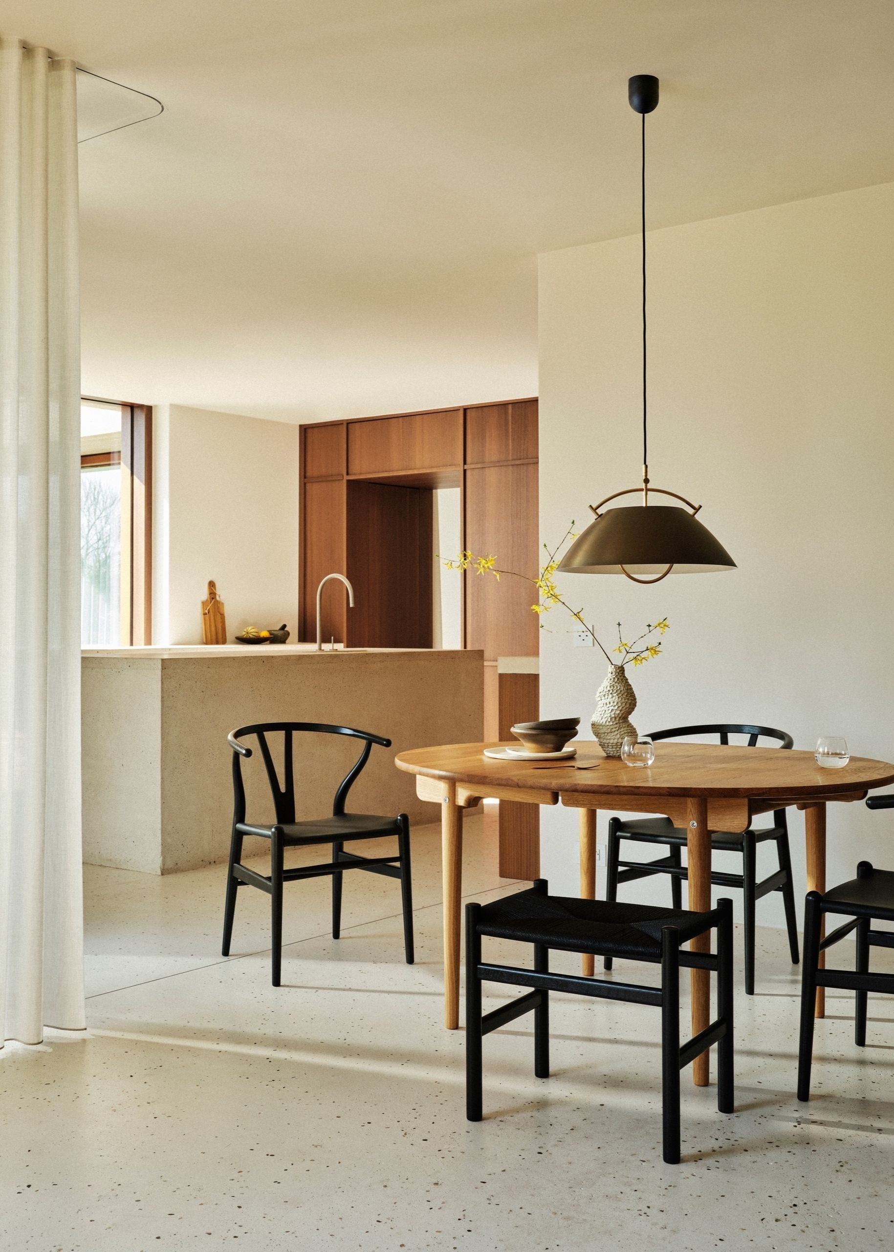 A bright, light kitchen with a large island, wooden cabinetry, and it opens into a dining space with a wooden table, and black dining chairs.