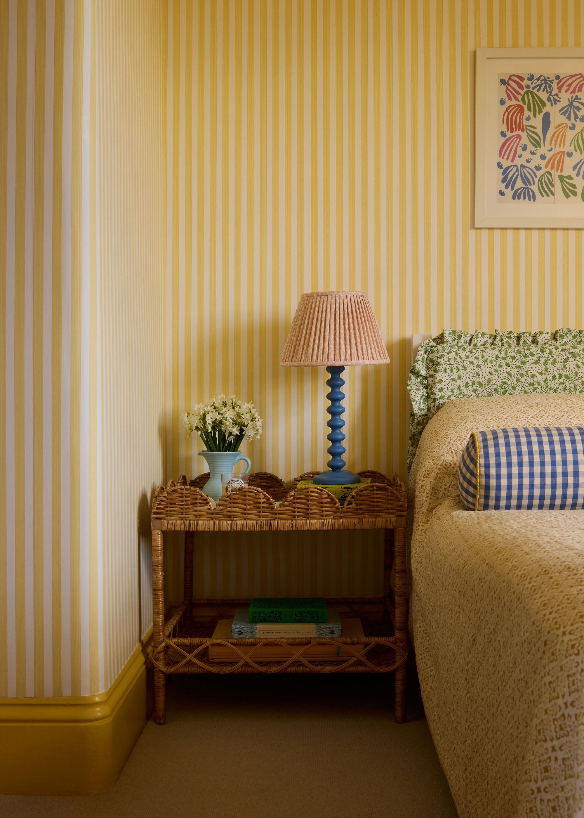 A cozy bedroom with yellow and cream striped wallpaper, dark yellow trim, yellow bedding with a green patterned headboard, and a rattan nightstand with a blue and red lamp.