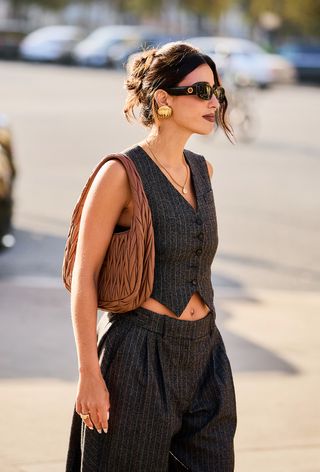 a close street-style photo of a woman wearing a miu miu bag with a pinstripe vest and matching pants and gold earrings