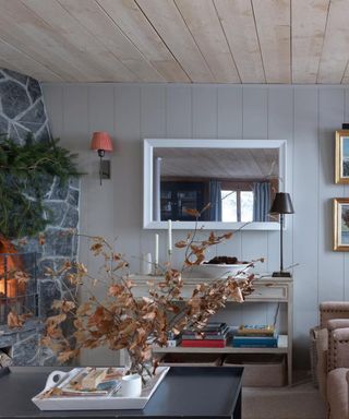winter decor ideas, cozy living room with grey shiplapped walls, stone fireplace lit, pine fir hung above, pair of tan upholstered armchairs, console, wall lights, coffee table with autumnal foliage in vase