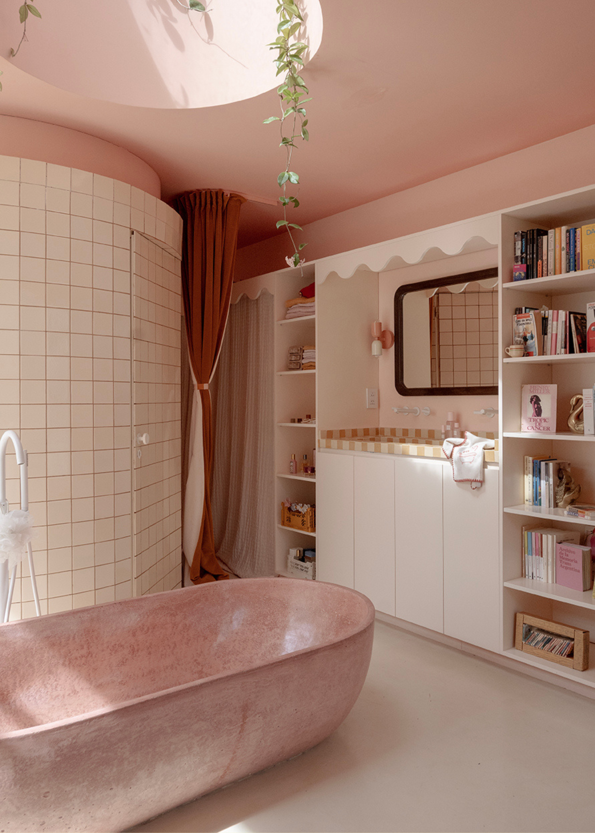 A pale pink tub in a bathroom with a skylight, beside white shelves of books and bath products, with a checked tile sink and curved mirror