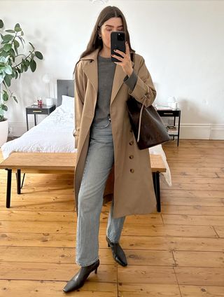 British fashion influencer Anna Newton poses for a mirror selfie wearing a trench coat, gray sweater, straight-leg jeans, and square-toe kitten heel boots.