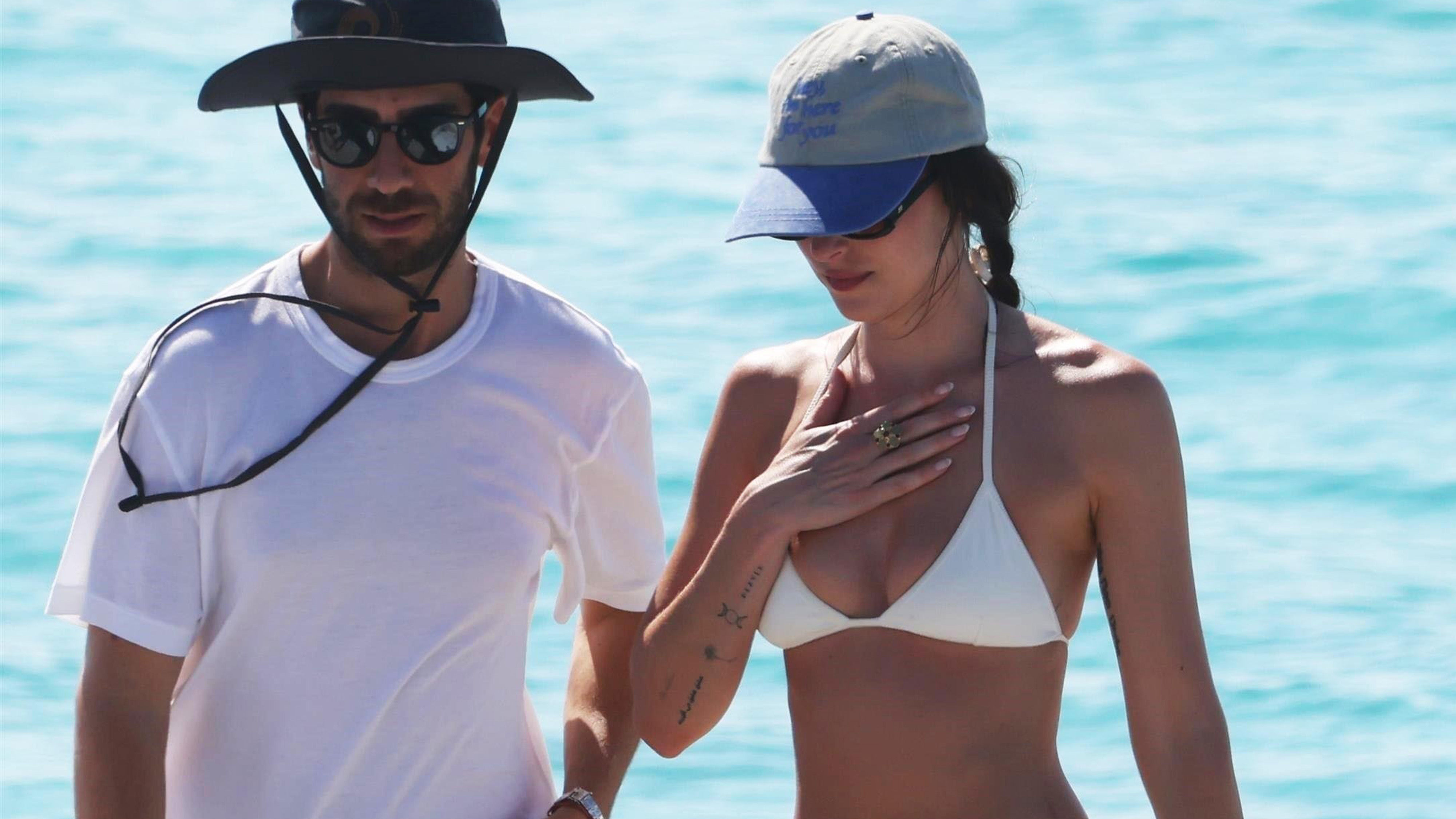 Dakota Johnson stands next to a man wearing a baseball cap, white bikini, floral skirt, and mesh flats.