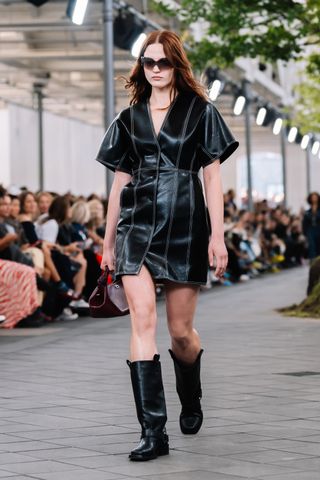 a model wears a black leather dress with mid shaft moto boots while walking down the Ganni runway