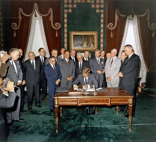 President Kennedy signs the Limited Nuclear Test Ban Treaty in the Treaty Room at the White House. 7th October 1963. He sits at a desk signing the treaty and is surrounded by men in suits.