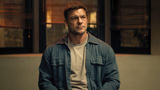 Alan Ritchson as Jack Reacher sitting at a table and looking to his right. 