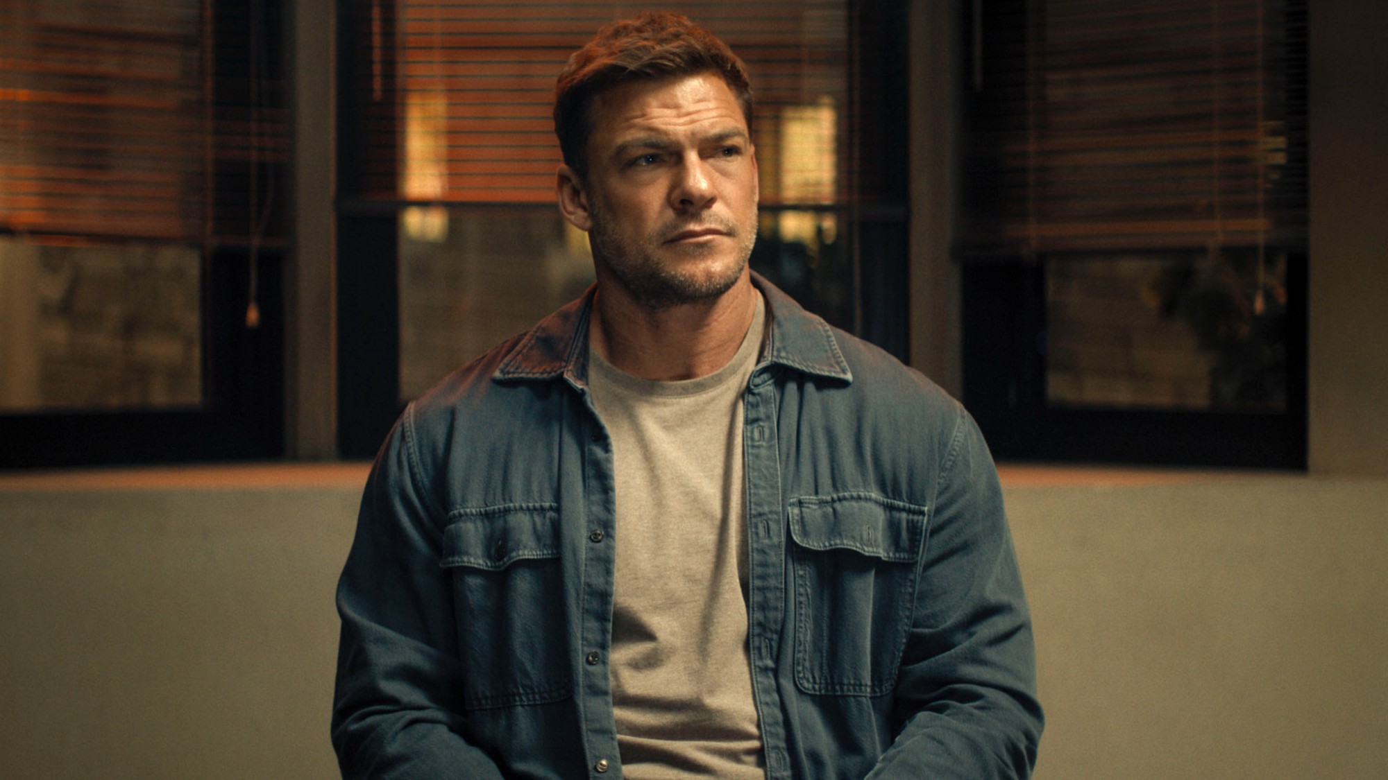 Alan Ritchson as Jack Reacher sitting at a table and looking to his right. 
