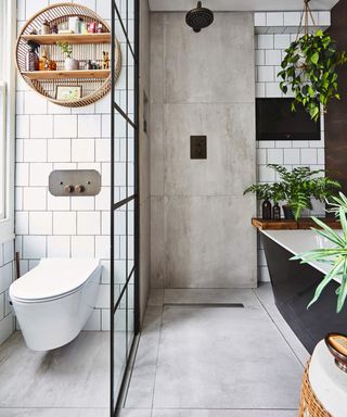 Bathroom with white toilet with white tiles over and rattan cylindrical shelf, concrete gray tiled shower and black framed shower screen and freestanding bathtub with black surround with plants around it