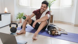 Man sits on exercise mat looking at open laptop, a kettlebell is next to him