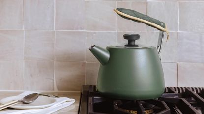 A green tea kettle on a gas stove, against a pink rustic tile backsplash
