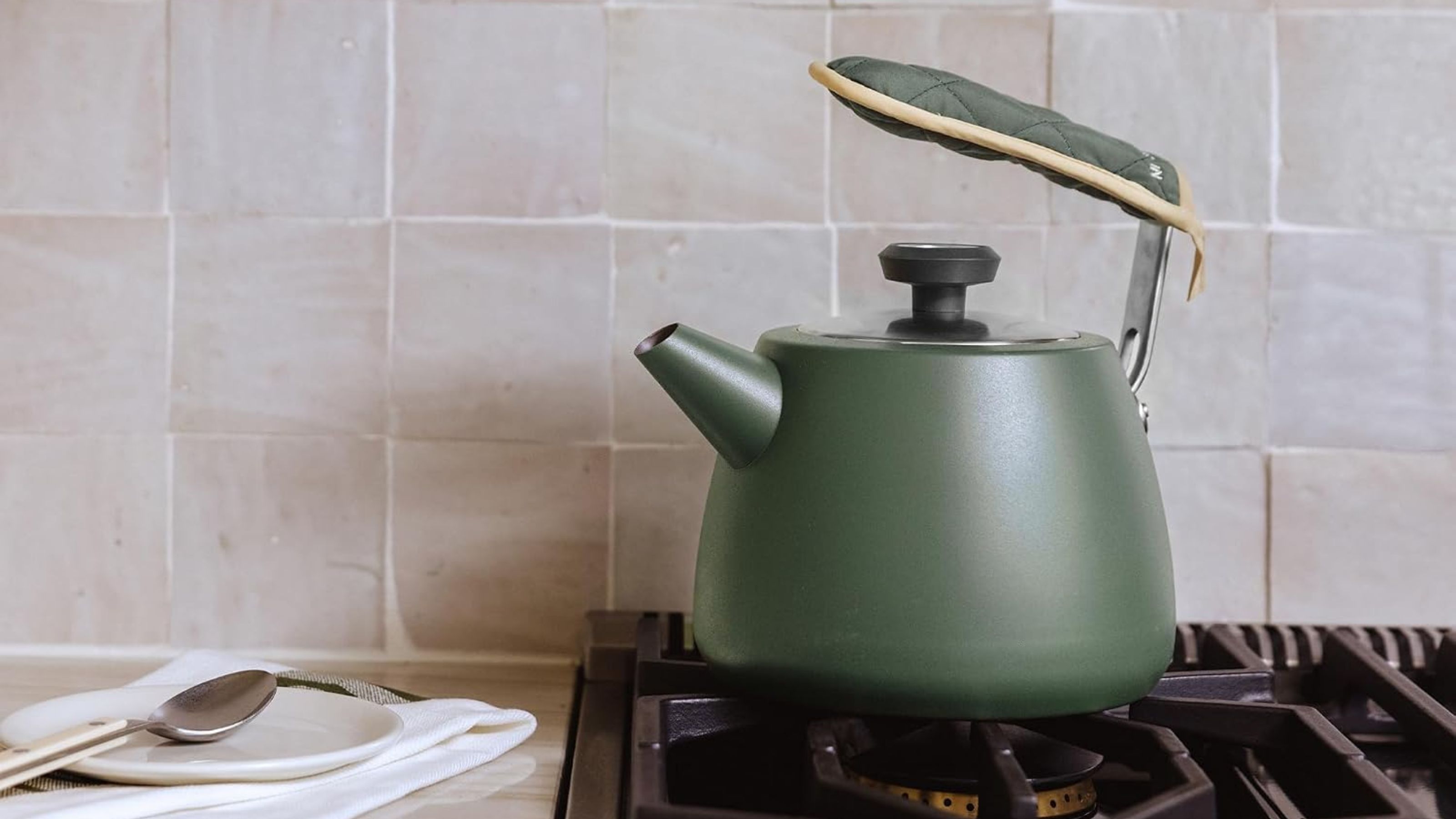 A green tea kettle on a gas stove, against a pink rustic tile backsplash
