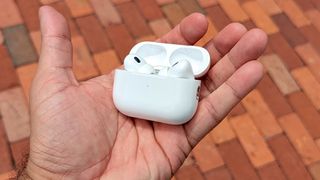 AirPods Pro 2 in charging case in reviewer's hand with red brick paving in background