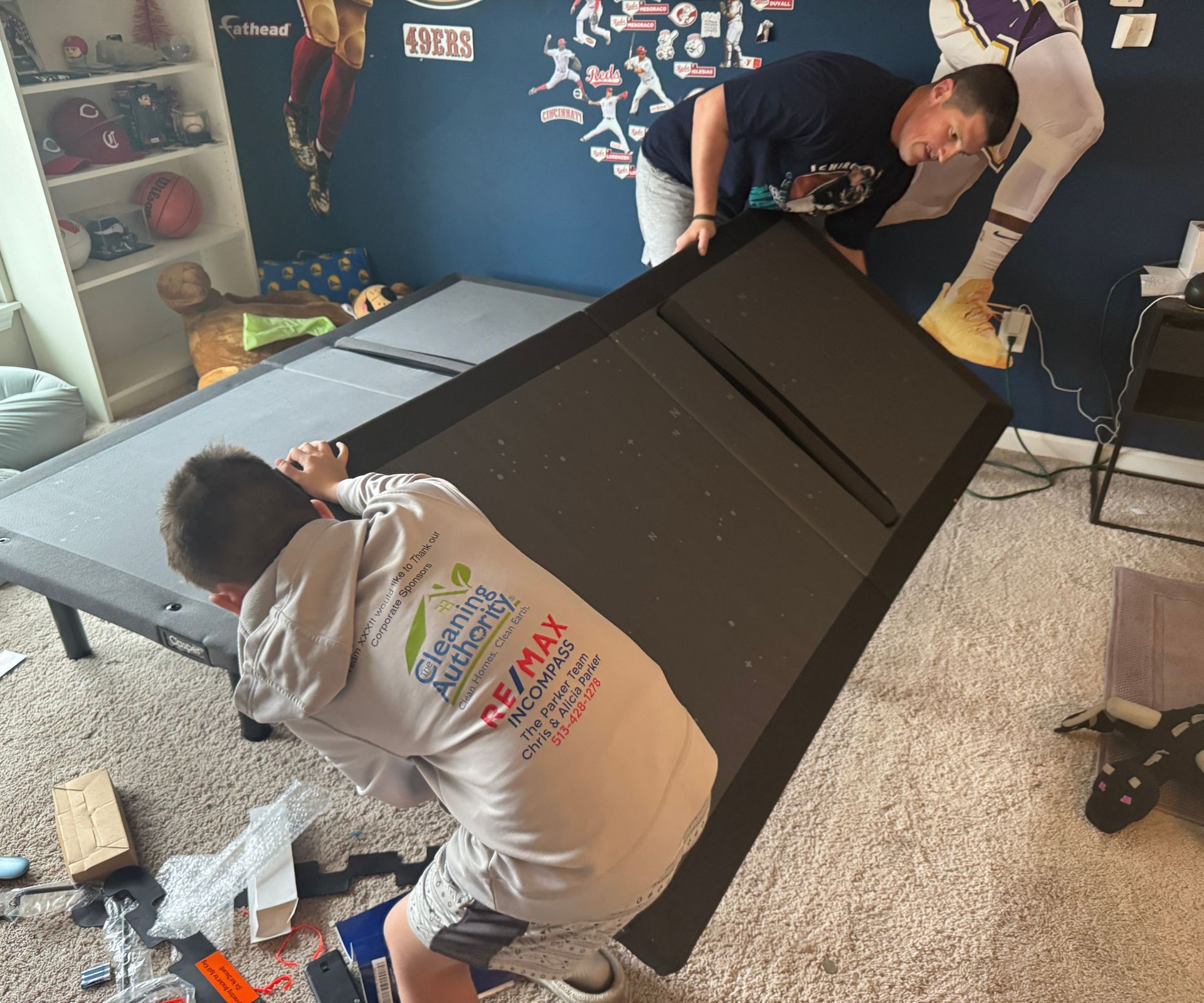 A man and a boy setting up the Casper Adjustable Base on a beige carpet against dark blue walls.