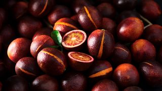 A dark, moody close-up of a pile of blood oranges, with a few cut open to reveal the striking red and orange interior.