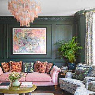 dark green colour drenched painted living room, with bold artwork on the wall, a pink velvert sofa and a patterned armchair and a pink glass chandelier