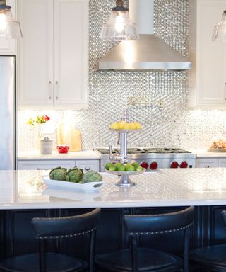 A white kitchen with a silver metallic splashback, white cabinets, a silver oven and hood, and a white countertop with a white plate of artichokes and a metal stand with limes and lemons, with navy blue seats in front