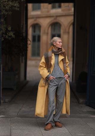 Amalie Gassmann wearing a yellow long coat, a colorful scarf, a grey sweater, grey pants and UGG chestnut Classic Mini II Boot on March 04, 2022 in Paris, France.