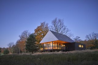Exterior view of an illuminated cabin by MacKay-Lyons Sweetapple Architects on Bigwin Island in the evening. The cabin features floor-to-ceiling windows, a pitched roof and it is surrounded by greenery