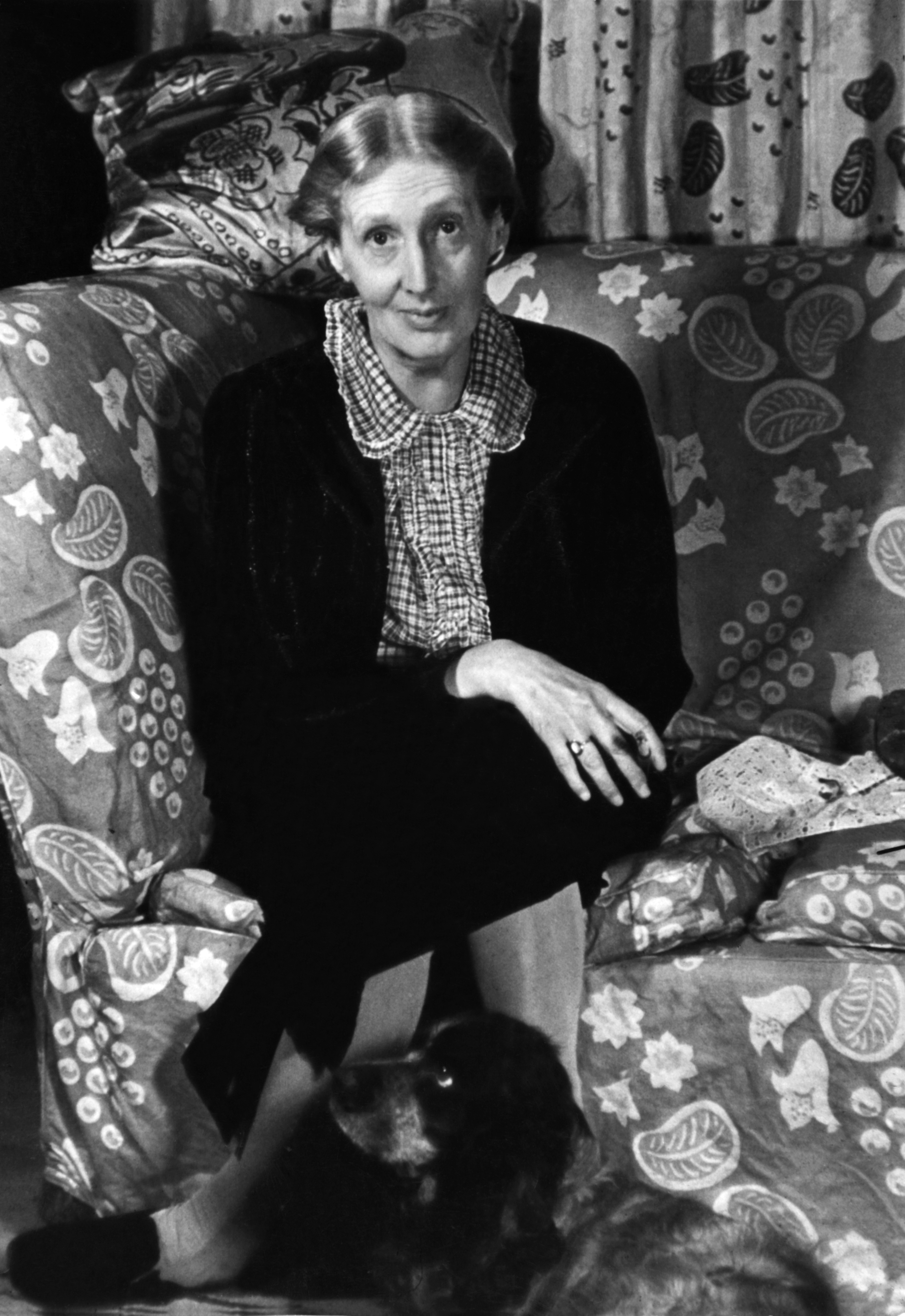 Portrait of English author Virginia Woolf sitting cross-legged on a couch, with her Cocker Spaniel, Pinka, at her feet, taken in London in 1939.