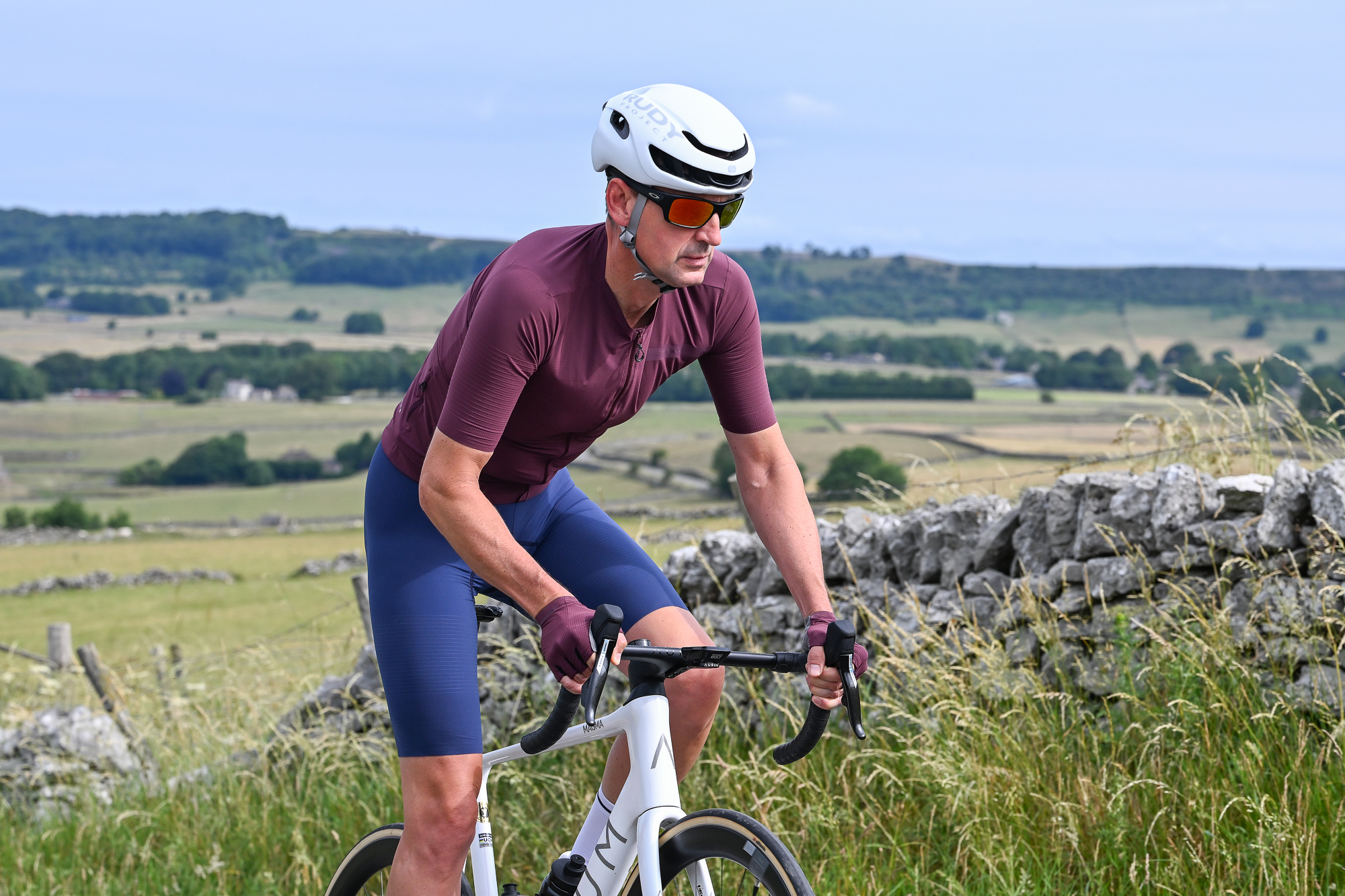 Man riding a white bike wearing a purple jersey, purple gloves, blue shorts and a white helmet