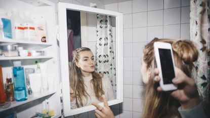 Cropped hand of woman photographing female friend in mirror reflection at dorm bathroom