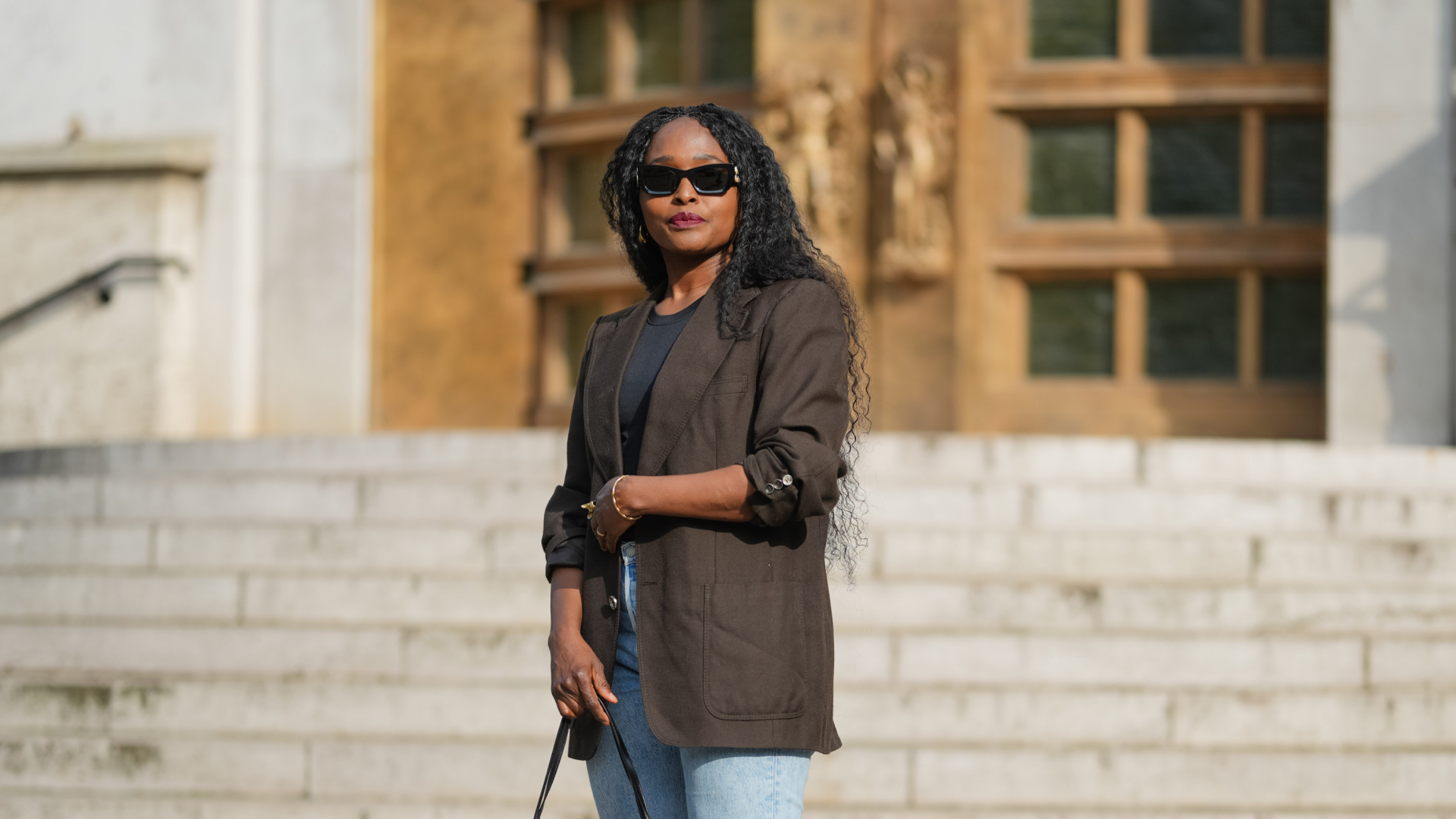 A woman wearing red lipstick, a blazer, and jeans, with goddess braids in her hair.