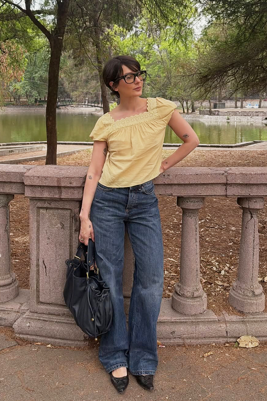 a woman wearing a yellow prairie top with blue baggy jeans, black pointed toe shoes, and a black leather tote bag