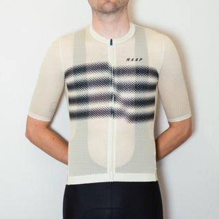 A white man stands against a white wall wearing black cycling shorts and a tight beige mesh jersey