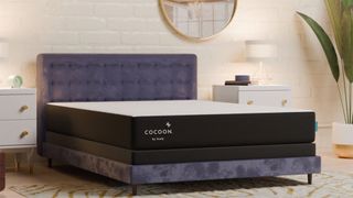 The black and white Cocoon by Sealy Chill mattress placed on a purple bed frame in a bedroom with a white brick wall, two bedside cabinets and a plant