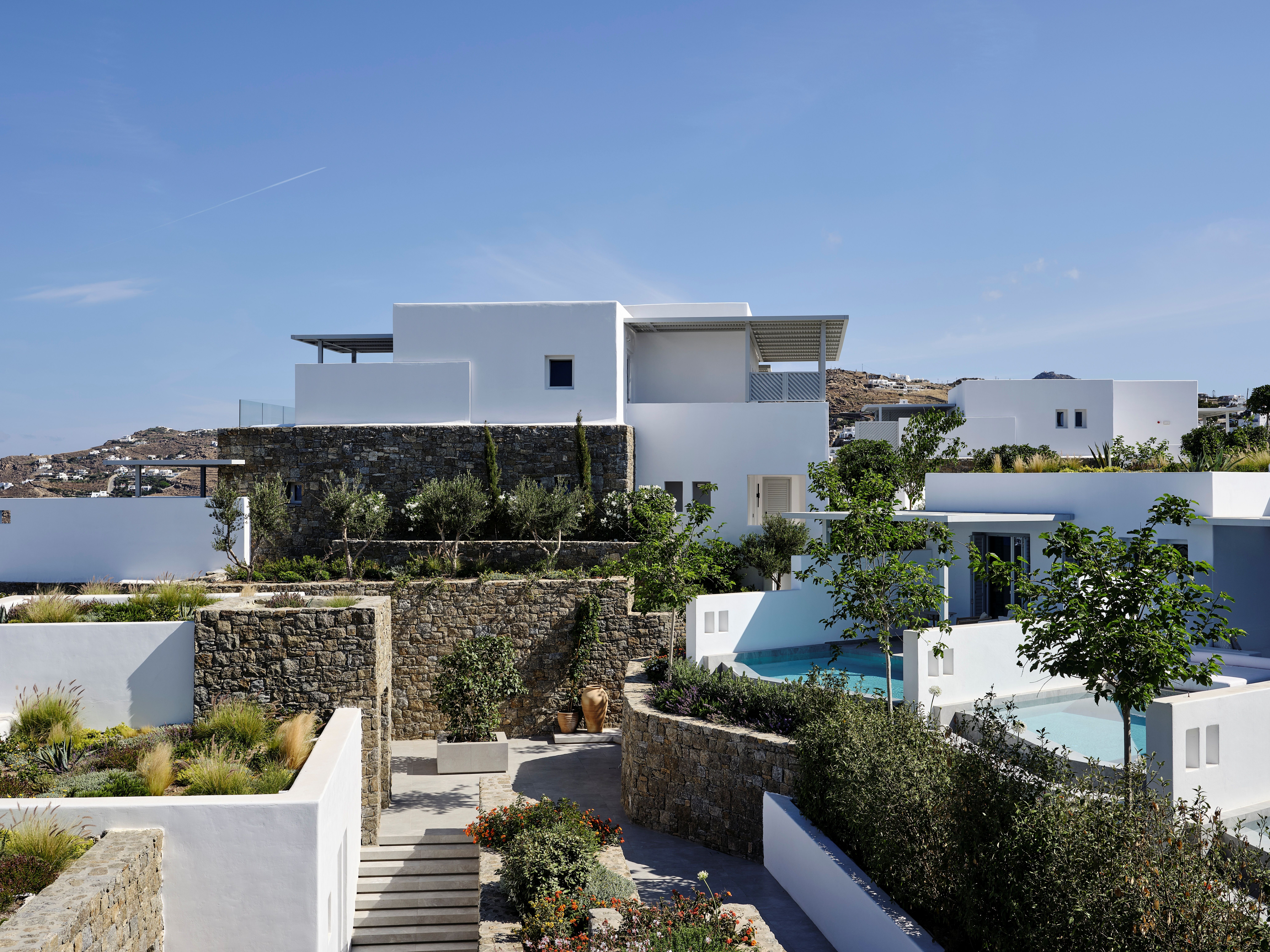 The exterior of a Cycladic-style luxury resort, with white-washed architectural volumes brought to life by thriving plants and individual swimming pools.