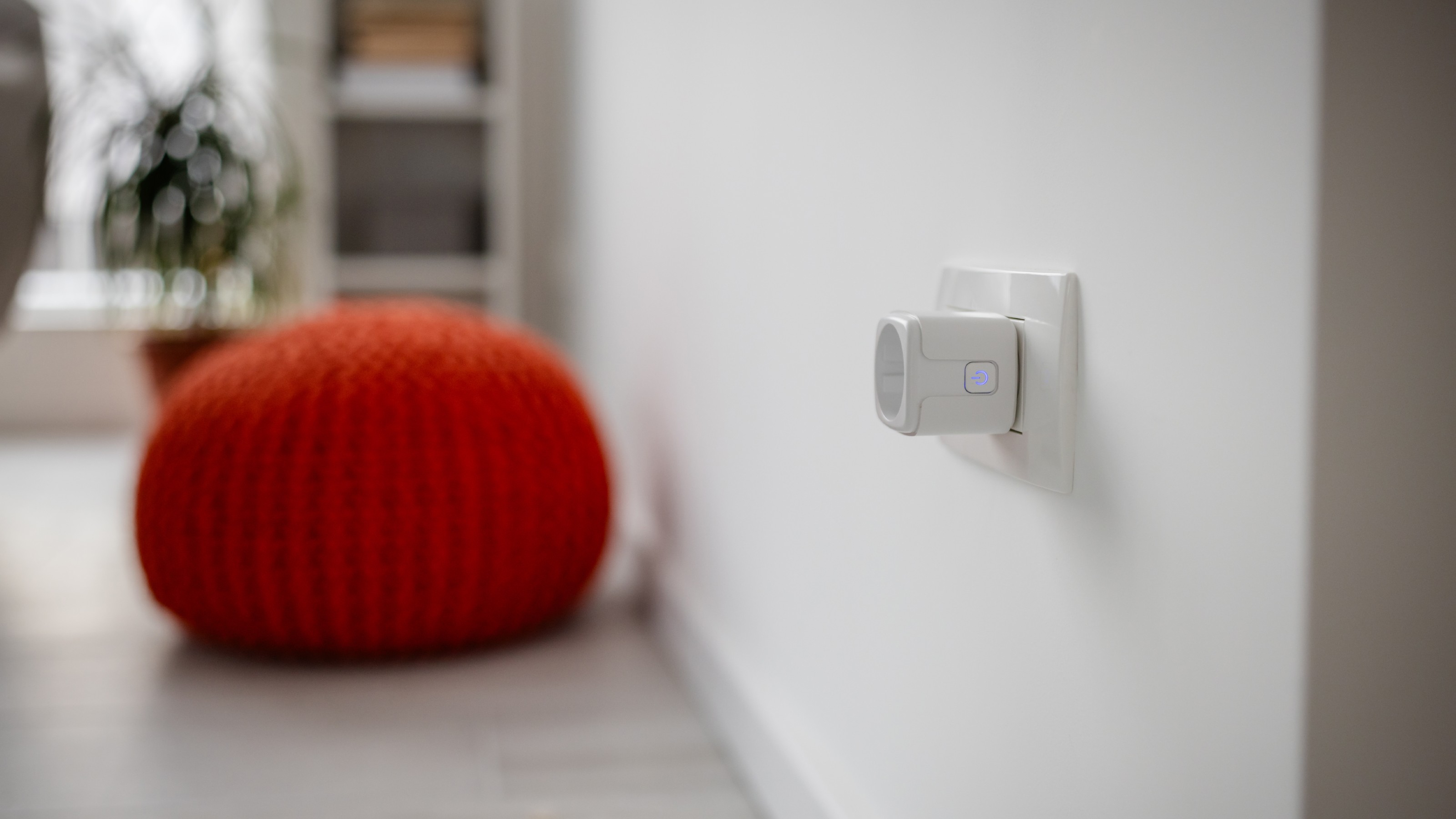 Smart plug in wall socket, with a burnt orange knitted pouffe and a plant in the background