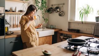 Woman drinking orange juice and scrolling on phone in kitchen, leaning against the counter