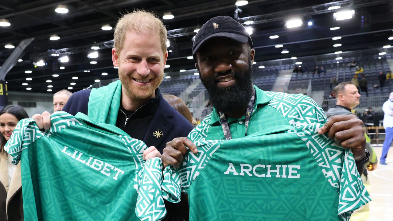 Prince Harry and a man smiling and holding green jerseys that say Lilibet and Archie in a basketball arena