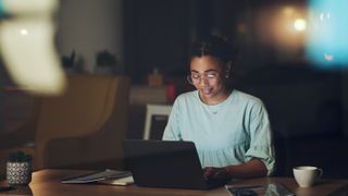 A woman sitting at a table and looking at a laptop, semingly reading, at night time