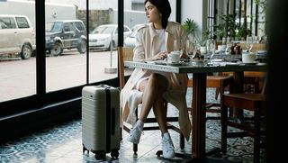 A female sat in a restaurant with a grey suitcase next to her.