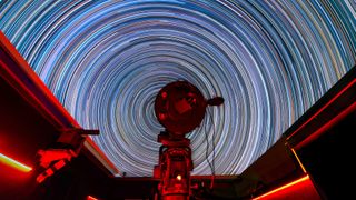 in the foreground, a telescope can be seen. in the background, stars turn into giant rings of different colors in the sky thanks to a long exposure photograph