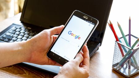 Google Chrome search page on a mobile phone being held in someone's hands at a desk with a laptop in the background