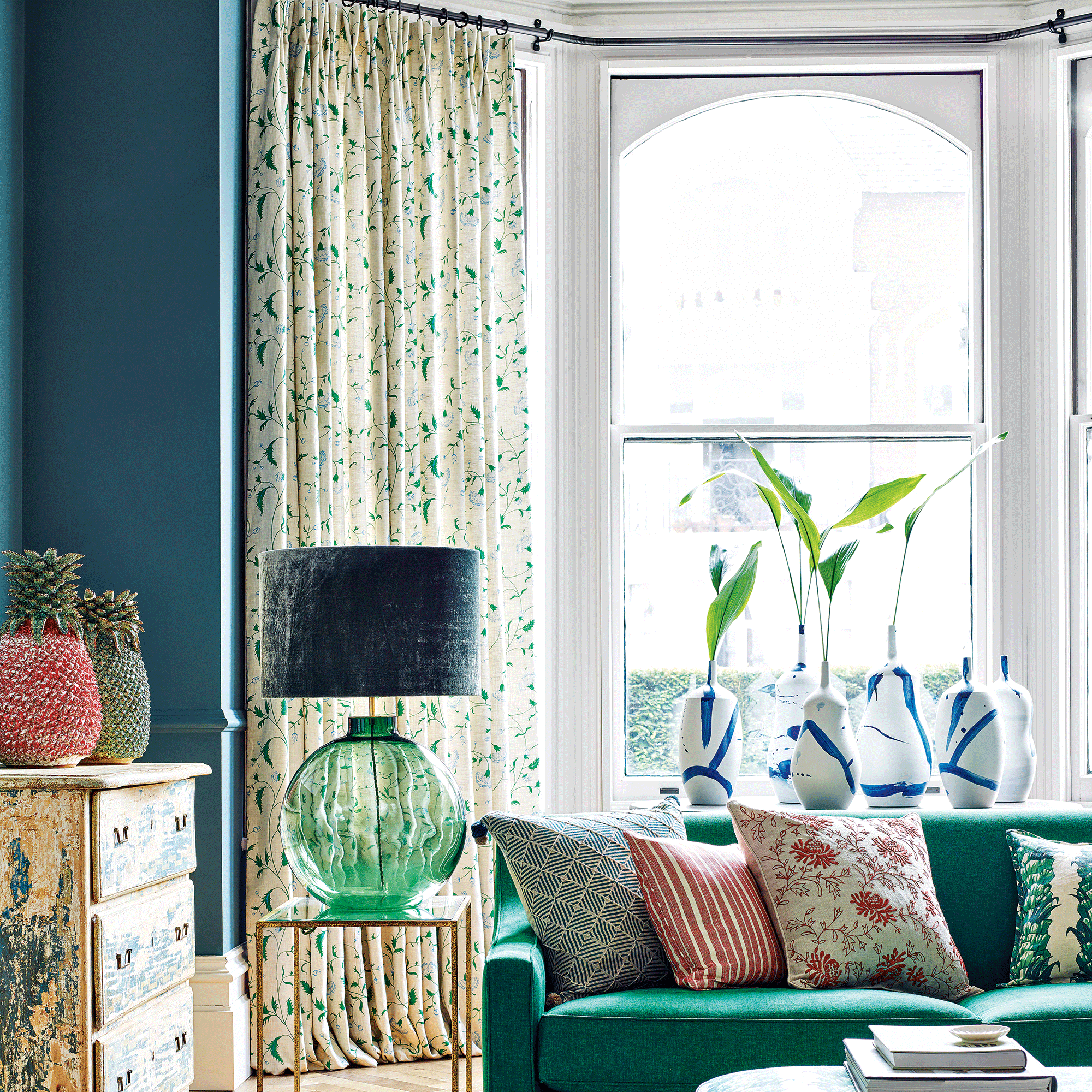 Navy blue living room with large sash bay windows and patterned curtains. A large green lamp and a green sofa sit infront of the window