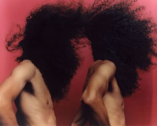 Photo of two shirtless people with their curly, wind blown hair covering their faces against a pink background