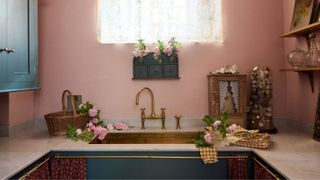 A romantic pink utility room with a brass sink, antique-style brass tap, and deep teal lower cabinets. Lace curtains, fresh flowers in jars, and vintage curios give the space a whimsical, nostalgic charm.