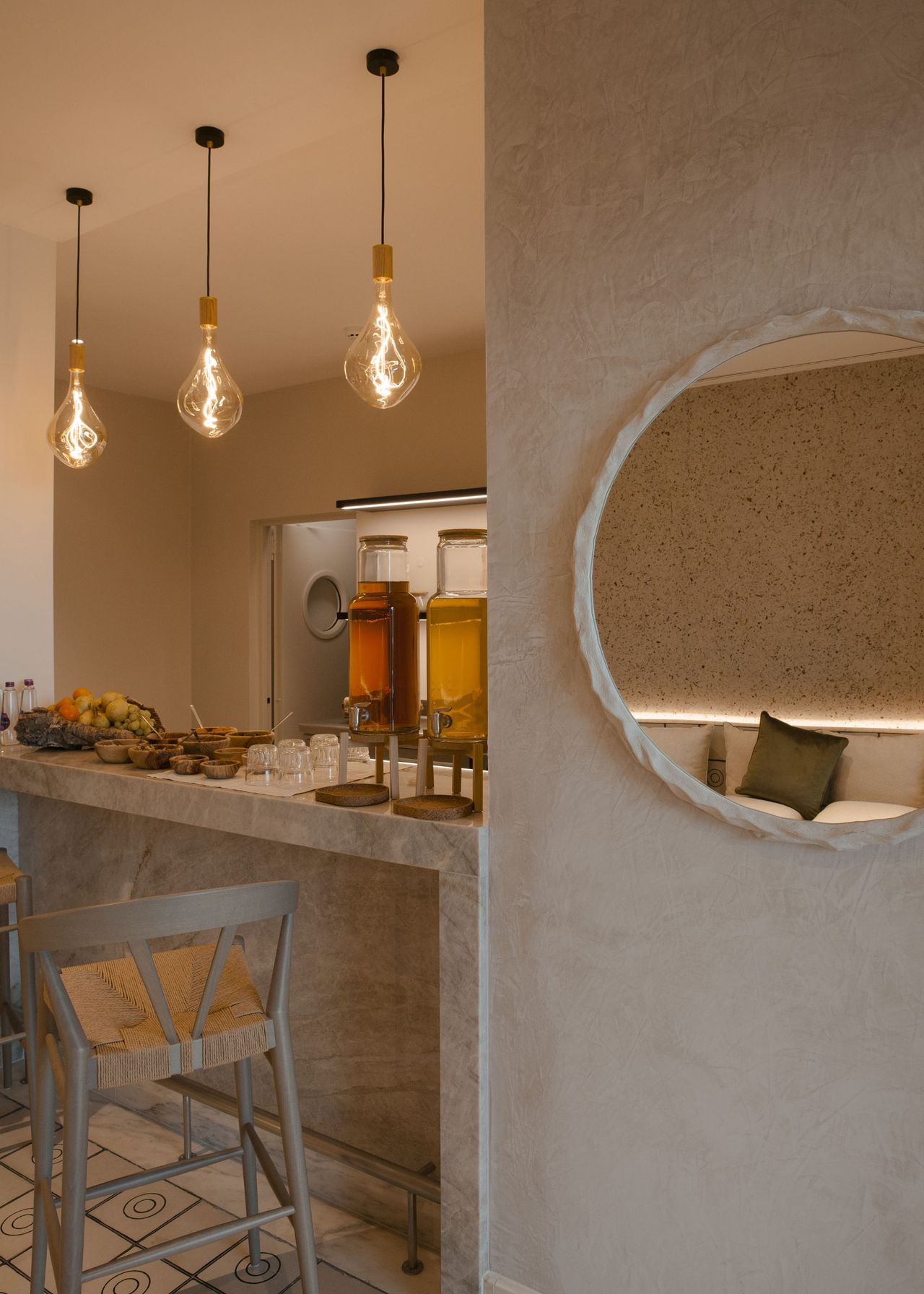 A bar stool beside a table with food and drinks. Three pendant lights are hanging above the table, as well as a large mirror on the side of the wall.