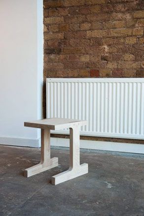 two legged wooden stooll photographed on a concrete ground with a brick wall and heating panel in the background