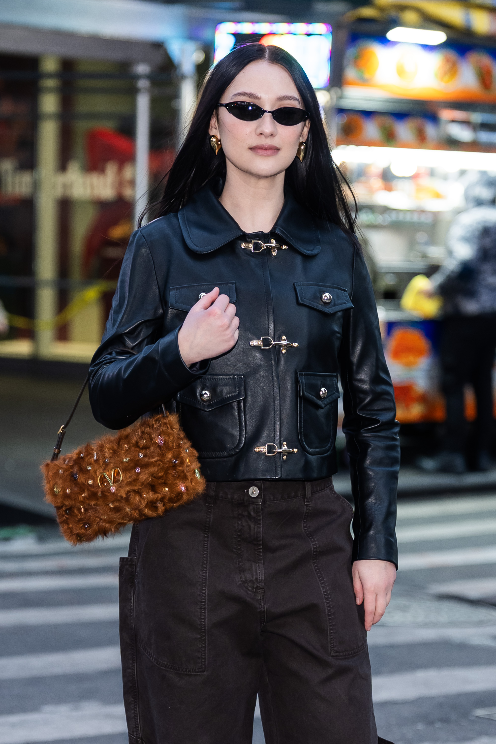 a paparazzi of a girl with long, dark hair standing in the middle of a crosswalk wearing a black leather jacket and brown pants