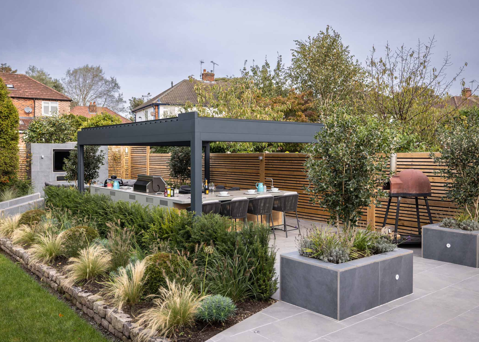 a sleek, minimal outdoor kitchen under a black pergola with a free-standing pizza oven and lovely planting around