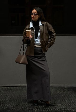 A black turtleneck outfit is shown in a photo of a woman standing on the sidewalk wearing a brown bomber jacket layered over a white button-down shirt over a black turtleneck sweater styled with a white belt, gray maxi skirt, ankle boots, and a brown shoulder bag