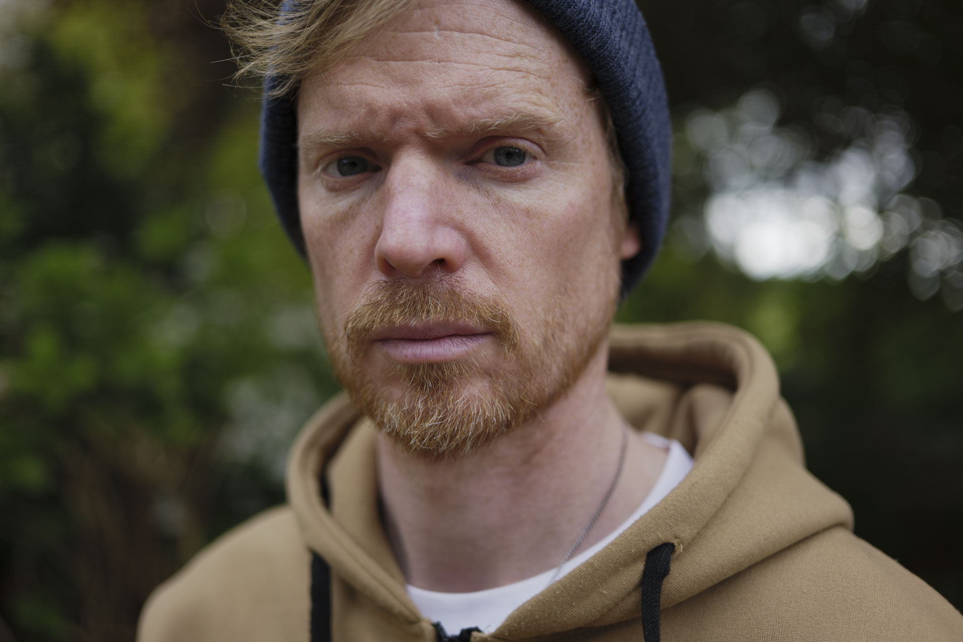 Portrait of a man in a beanie hat and hoodie, the background is lush green bushes and trees with dappled light