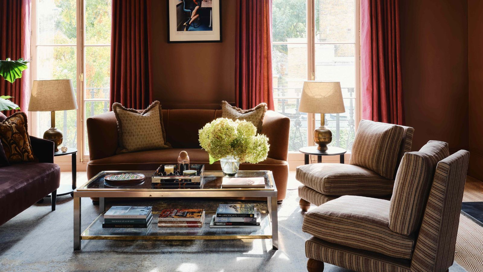 dark orange brown living room with orange curtains, large windows, a tan sofa, striped slipper chairs with bun feet and a glass coffee table