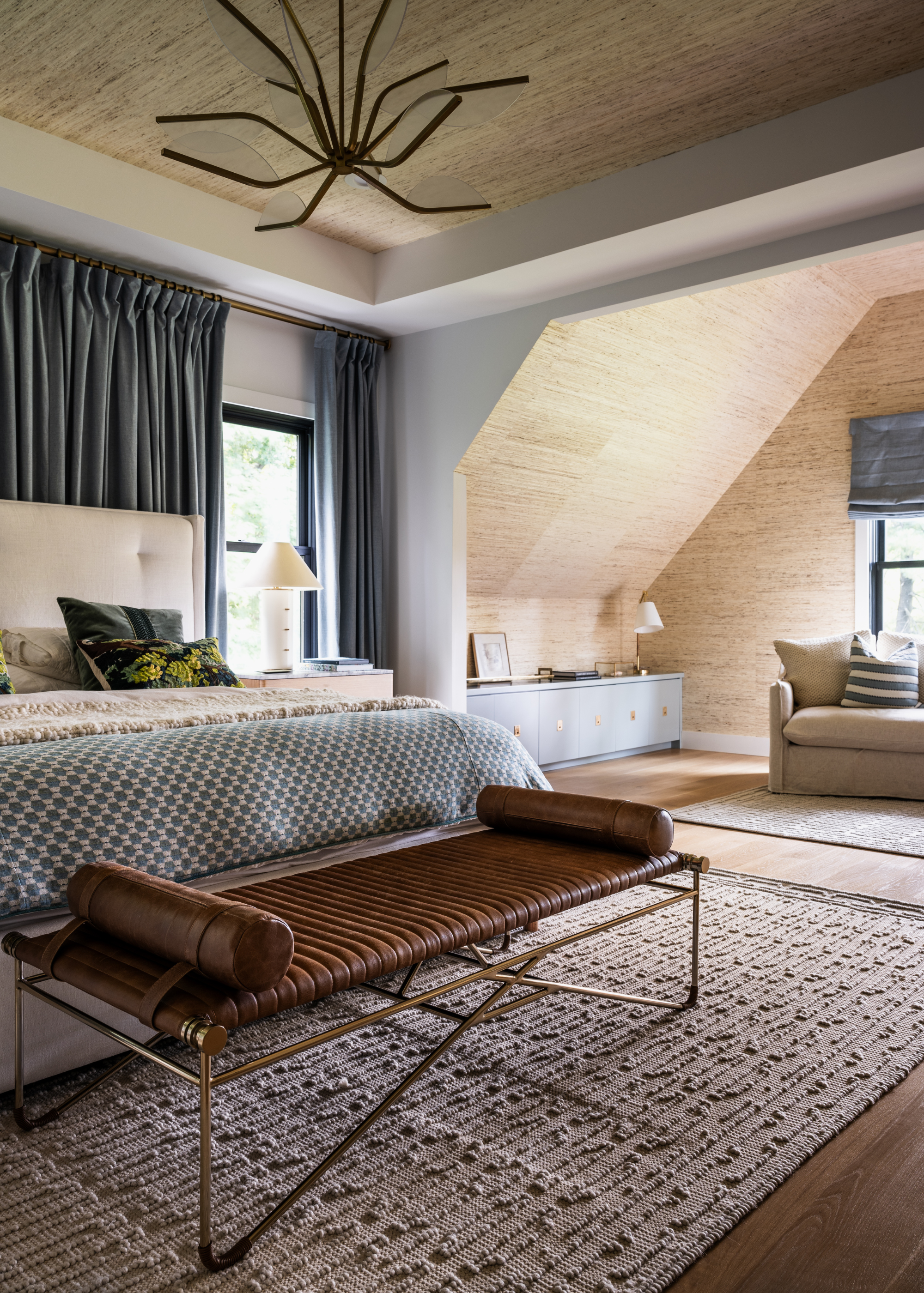 A bedroom with a tubular pendant light, a textured area rug, a leather bench, a patterned bedscape, and a sofa