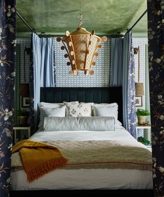 A bedroom with a green ceiling, a sculptural pendant light, patterned blue and white wallpaper, and patterned drapes hanging around the bed